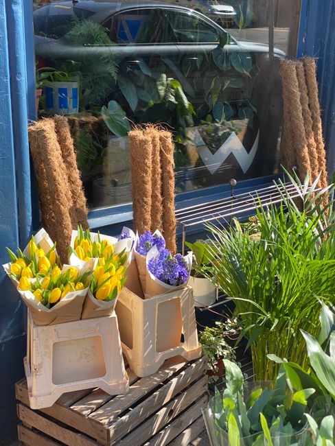 A display of freshly cut flower bouquets outside a floral shop, featuring a variety of flowers including bright yellow tulips and purple lavender arranged in brown wrapping paper. The bouquets are pla