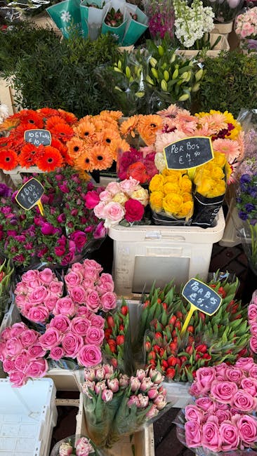 A vibrant display of fresh flower arrangements at a florist stall, featuring bright orange gerbera daisies, soft pink roses, yellow carnations, and red-tipped tulips. The flowers are grouped in white 
