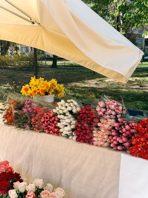 A vibrant display of freshly cut flower bouquets arranged on a market stall table under a large cream-colored umbrella. The selection includes tulips in shades of pink, white, and red, tightly grouped