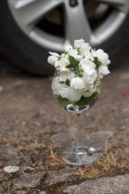 A floral arrangement of fresh white bell-shaped flowers, possibly lilies of the valley or similar, placed in a clear glass goblet-style vase. The bouquet features lush green leaves and delicate petals