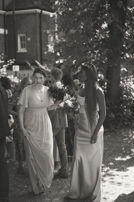A black and white photograph capturing a floral arrangement scene near the William Morris Gallery Walthamstow. In the foreground, two women dressed in elegant, flowing dresses are holding bouquets of 