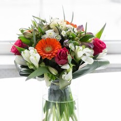 This lavish bouquet echoes the artistic pulse of Walthamstow-where tradition and innovation blend in every thoughtful detail. Large gerberas, glowing in shades of fiery orange and creamy peach, greet the viewer with the same warmth found among neighbours near Lloyd Park. Deep fuchsia roses add an elegant, romantic anchor, while pristine white alstroemeria and dainty clusters of spray roses lend airiness and subtle contrast. Tucked amid the flowers, broad aspidistra leaves are expertly curved and layered to form a protective, verdant frame, grounding the bouquet's vibrant colours. Smaller pointed leaves and trails of greenery weave between the blooms, adding fullness and a sense of lush abundance as seen in nearby gardens. Housed in a tall, crystal-clear glass vase, the arrangement's green stems create a living sculpture beneath the blossoms. The natural lighting highlights every hue and texture, evoking feelings of optimism and joy-perfect for celebrating life's milestones, sending gratitude, or simply adorning a dining table after a day at the William Morris Gallery. This composition is a testament to the talented florists at Flower Delivery Walthamstow and the neighbourhood's welcoming character.